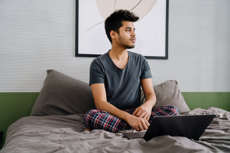 Young indian thoughtful man with laptop sitting on bed in cozy room at homeの写真素材