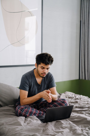 Young handsome indian man sitting on bed with folded hands and looking at laptop in cozy bedroom at homeの写真素材