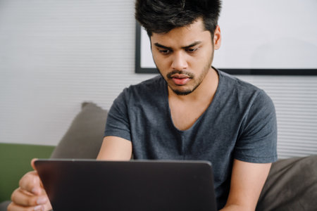 Young indian focused man looking on laptop screen sitting on bed in cozy room at homeの写真素材