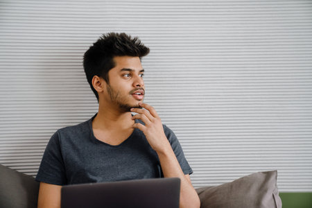 Young indian thoughtful man touching chin with his hand looking rightward sitting with laptop at...