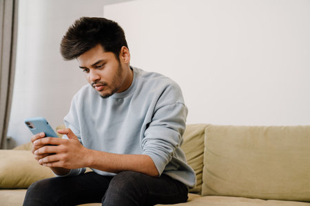 Young indian man sitting on sofa and scrolling news on his phone at homeの写真素材