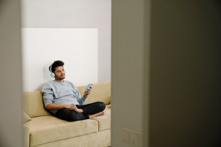 Young indian man in headphones with phone sitting on couch in lotus pose at homeの写真素材