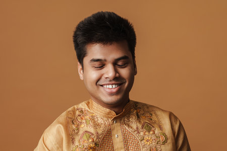 Young brunette indian man smiling at camera with eyes closed isolated over yellow background