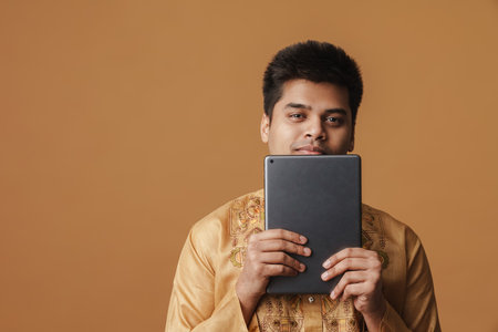 Young brunette indian man smiling and holding tablet computer isolated over yellow background