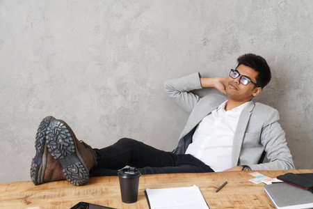 Young brunette indian businessman smiling and sitting at table in office over grey wallの写真素材