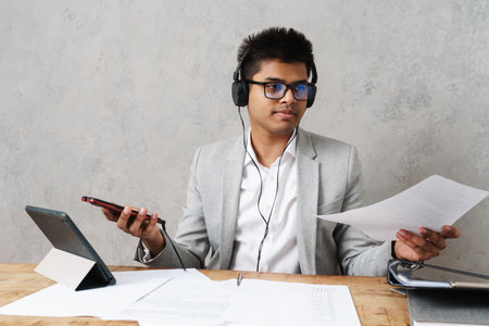 Young happy indian businessman using cellphone and headphones while working in office