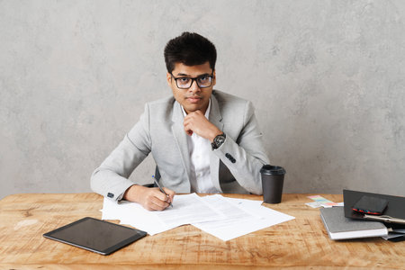 Young serious indian businessman working with paper documents while sitting at table in office