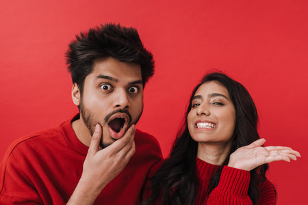 Young indian couple expressing surprise while posing together isolated over red background