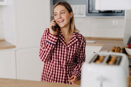 Young white woman wearing pajama standing by stove in kitchen and talking on cellphoneの写真素材