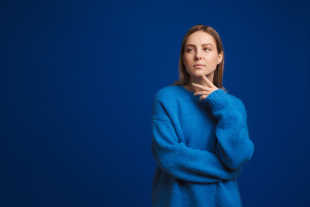 Young thoughtful beautiful girl touching chin and pondering looking leftward over blue isolated backgroundの写真素材