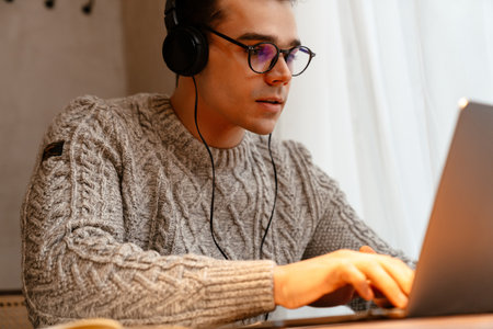 Young white man wearing eyeglasses smiling while using laptop and headphones in cafe