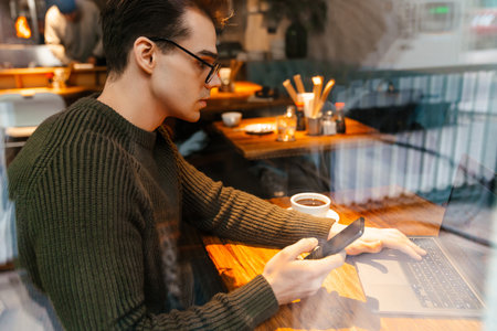 Young white man wearing eyeglasses using cellphone and laptop in cafe
