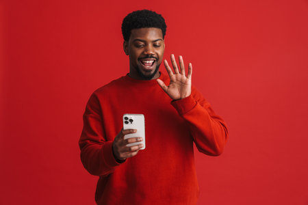 Black bearded young man smiling and using cellphone isolated over red backgroundの写真素材