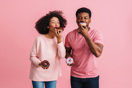 Black happy man and woman having fun and eating donuts isolated over pink background