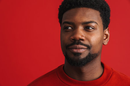 Black bearded young man smiling and looking aside isolated over red backgroundの写真素材