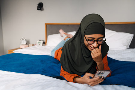 Young beautiful woman in hijab and glasses liying on the bed and looking on her phoneの写真素材