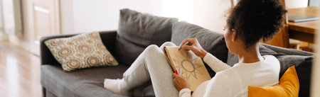 Backview of pregnant latin woman drawing flower with a pencil in a sketchbook sitting on sofa at homeの写真素材