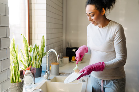 Adult beautiful pregnant african woman washing dishes in cozy sunny kitchen at homeの写真素材
