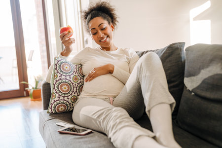 Adult beautiful pregnant smiling african woman eating apple and holding her belly while sitting on sofa at homeの写真素材