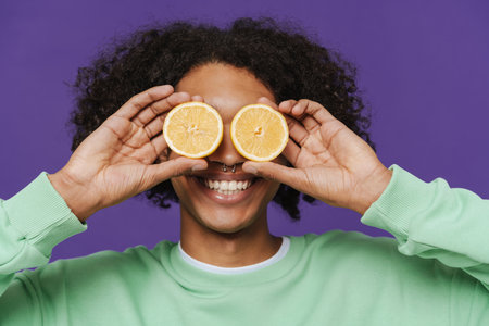 Young caribbean man smiling while making fun with lemon isolated over purple backgroundの写真素材