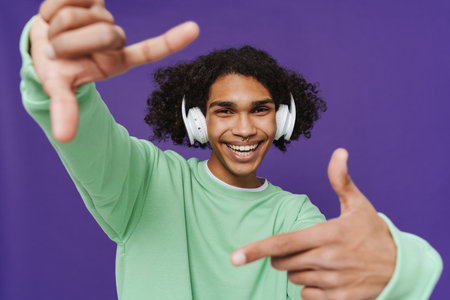 Young smiling happy curly latin man in headphones with piercing looking at camera and showing frame gesture while standing over isolated violet backgroundの写真素材