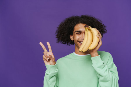 Young latin smiling curly man covering his face with bunch of bananas, showing victory gesture and looking at camera with one eye, while standing over isolated violet backgroundの写真素材