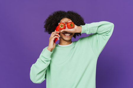 Black man talking on cellphone while making fun with tomatoes isolated over purple background