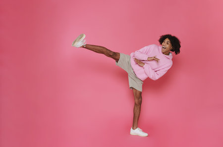 Young smiling handsome stylish latin curly man showing fighting pose with leg kick over isolated pink backgroundの写真素材