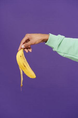 Male hand holding banana peel cropped close-up over isolated violet backgroundの写真素材