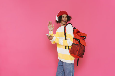Young caribbean man in headphones gesturing and using cellphone isolated over pink background