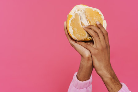 Black man's hands holding and showing pamelo at camera isolated over pink background