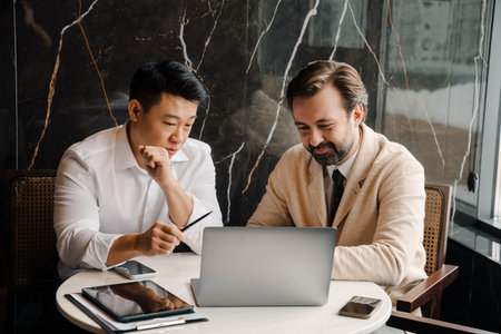 Adult multiracial businessmen talking and using laptop while sitting by table indoorsの写真素材