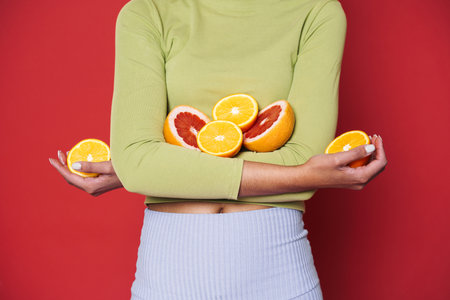 Young black woman with afro curls having fun holding oranges cut in half isolated over red...