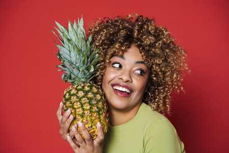Young black woman laughing while posing with pineapple isolated over red background