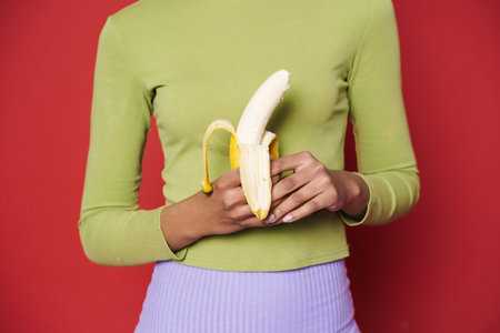 Young black woman wearing green sweater posing with banana isolated over red backgroundの写真素材