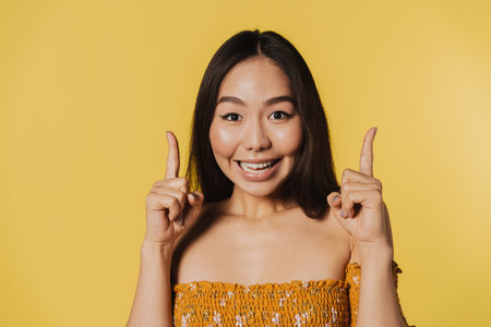 Young asian woman smiling while pointing fingers upward isolated over yellow backgroundの写真素材