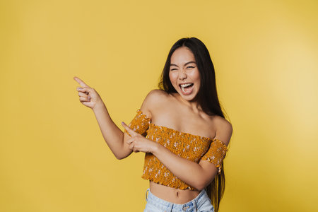 Young asian woman smiling while pointing fingers aside isolated over yellow backgroundの写真素材