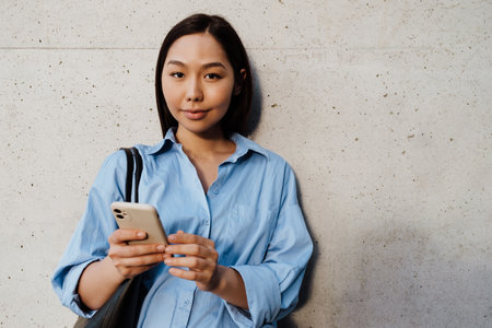 Asian brunette woman using mobile phone while standing by wall outdoorsの写真素材