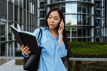 Asian brunette woman talking on mobile phone while standing with planner outdoorsの写真素材