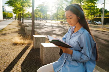 Asian brunette woman in headphones reading book while sitting on bench outdoorsの写真素材