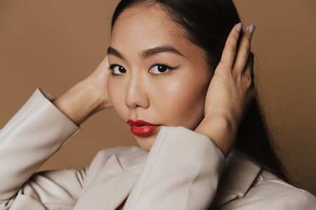 Young asian woman wearing jacket posing and looking at camera isolated over beige backgroundの写真素材