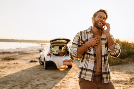 Young white man talking on cellphone while standing by car at seashoreの写真素材
