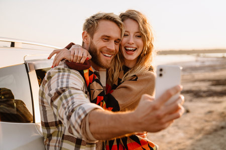 Happy young white couple taking selfie photo by car while walking at seashore on sunny dayの写真素材