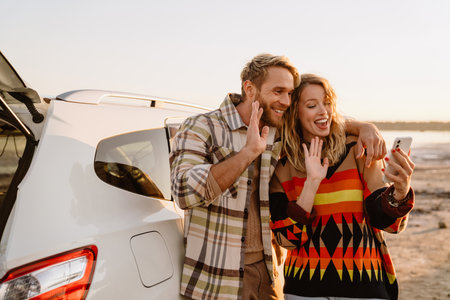 Happy young white couple taking selfie photo by car while walking at seashore on sunny dayの写真素材