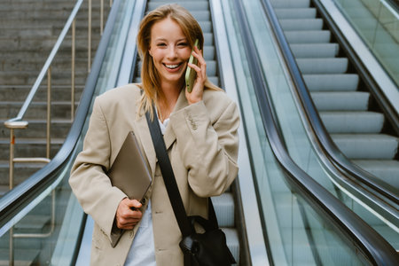 Young white woman talking on cellphone and holding laptop while going down escalator outdoors