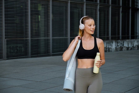 White young woman wearing headphones walking with water bottle after workout outdoors