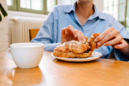 Young white businesswoman drinking coffee and eating croissant while sitting by table in cafeの写真素材