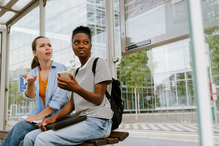 Young multiracial women using mobile phone together while sitting at bus station
