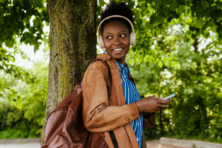 Young beautiful african smiling happy woman in headphones with phone looking back, while walking in parkの写真素材