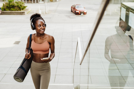 Young beautiful smiling fit african woman in headphones with phone and yoga mat looking aside, while walking stairs outdoorsの写真素材